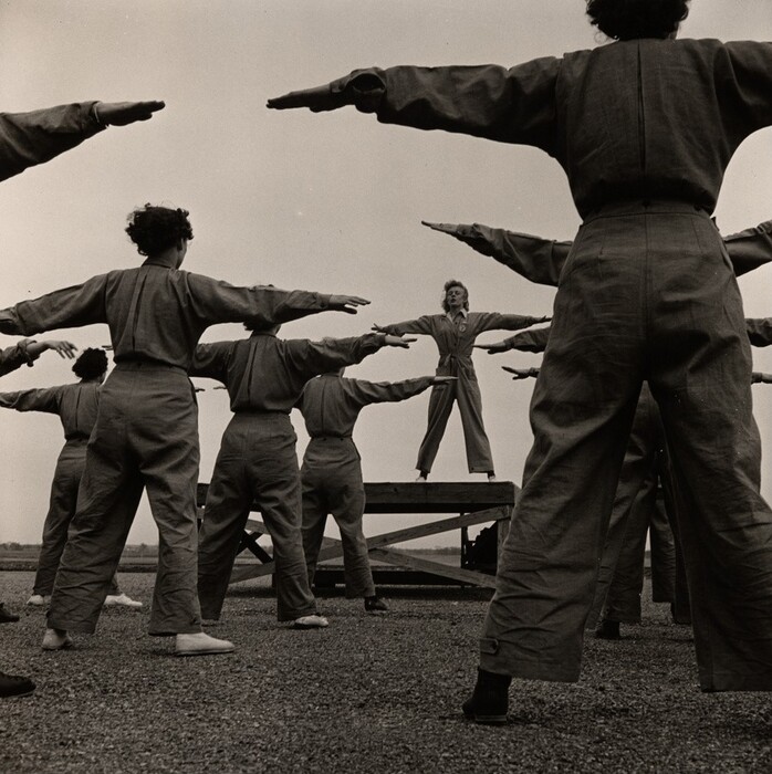 This is a photograph of a group engaged in an exercise routine. The image showcases several individuals wearing uniforms standing in a semi-circle with their arms raised and bent to the side in a synchronized exercise movement. In the center, elevated on a platform, another individual stands facing the others. The setting is outdoors, with a vast open space visible in the background, and the image is captured in black and white.
