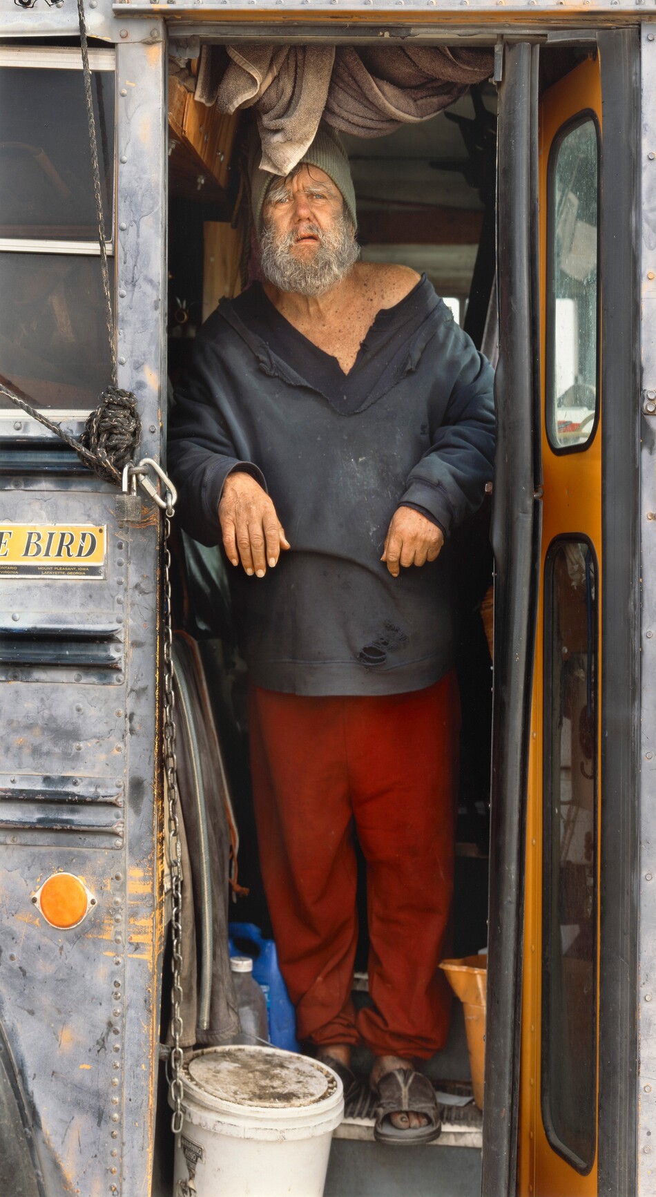 The image depicts a man standing in the doorway of a bus. He is leaning forward with both hands on the door frame. The man has a gray beard, weathered facial features, and light-colored eyes. He is wearing a beanie, a dark sweater, red pants, and open-toed slippers. Inside the bus are hanging towels, containers, and a white bucket near his feet. The interior is gray and yellow, while the exterior shows signs of rust and wear.