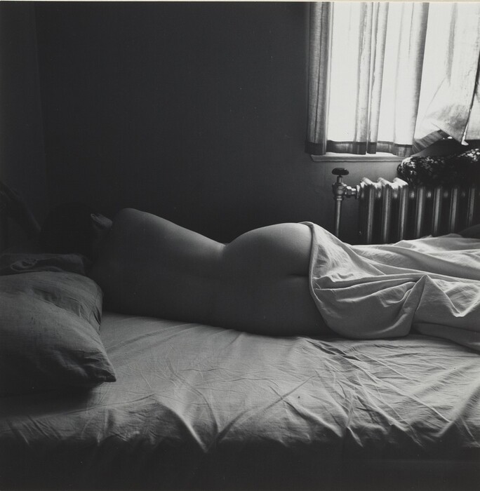 The image shows a person's silhouette lying across a bed, with their body mostly covered by a sheet except for parts of their back and buttocks. The person's head is turned away from the camera, resting on a pillow, so their facial features are not visible. Their hair appears to be dark and short. They do not appear to be wearing any clothing that is visible. The bed is situated near a window with light streaming in behind drawn curtains, casting a soft glow in contrast to the otherwise dimly lit room. A radiator is visible beside the window.