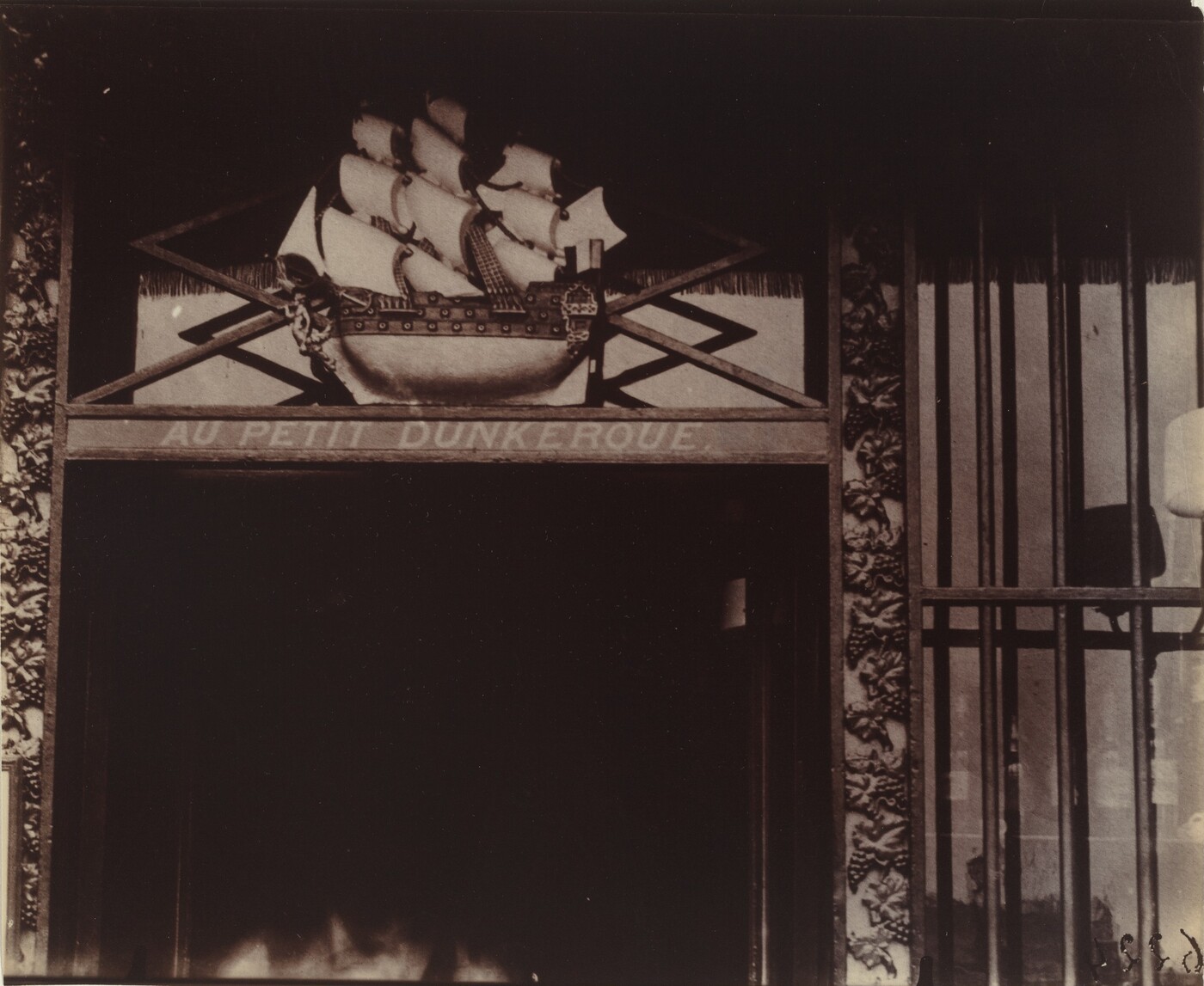 This is a photograph of a detailed architectural feature above an entrance. The image captures a wooden facade with a relief sculpture of a sailing ship with multiple sails. Below the ship is a horizontal panel with the inscription "AU PETIT DUNKERQUE." Additional ornaments with carvings of leaves and floral motifs surround the sculpture. Vertical bars or columns are visible to the right. Rich textures and shadows enhance the architectural elements.
