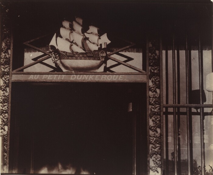 This is a photograph of a detailed architectural feature above an entrance. The image captures a wooden facade with a relief sculpture of a sailing ship with multiple sails. Below the ship is a horizontal panel with the inscription "AU PETIT DUNKERQUE." Additional ornaments with carvings of leaves and floral motifs surround the sculpture. Vertical bars or columns are visible to the right. Rich textures and shadows enhance the architectural elements.