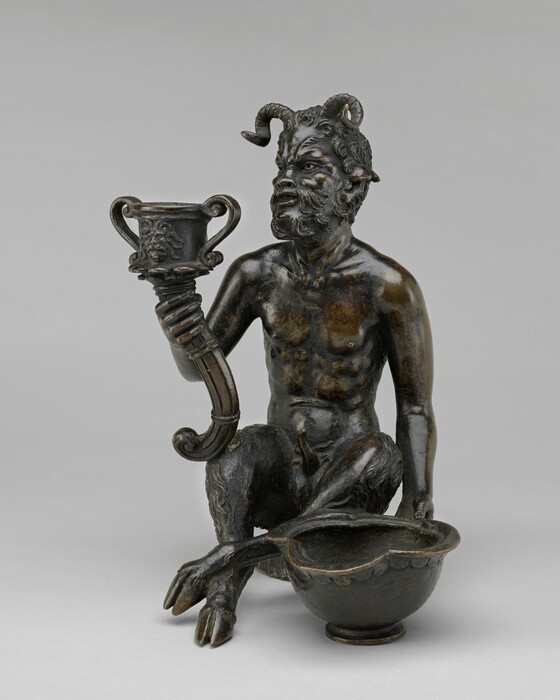 The sculpture depicts a mythological figure resembling a satyr or faun. It has a human-like upper body and the legs of a goat with hooves. The figure has a bushy beard, elongated pointed ears, and two curled horns on its head. It holds a drinking vessel in its right hand and a bowl-like vessel in its left hand. The sculpture is a deep brownish hue with a smooth finish, suggesting it may be made of bronze. The creature is seated with one knee up. The detailing adds depth to the sculpture, including textures in the hair, beard, and legs. The figure is looking slightly upwards.