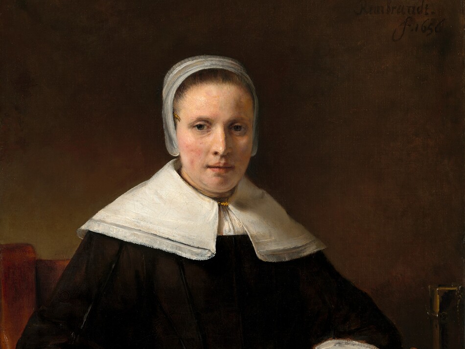 Shown from the lap up against a dark background, a pale-skinned woman wearing a black dress looks out at us in this vertical portrait. Soft light coming from our left highlights her oval face, the peach blush on her cheeks, and her crisp white collar, cap, and cuffs. Her brown hair is pulled back under the cap that flares slightly over her ears and the wide, lace-trimmed collar covers her shoulders. She has dark eyes, a straight nose, and her pink lips are slightly parted. Her left arm, on our right, rests on a table covered with a rose-red patterned carpet next to two pieces of fruit, perhaps apples, and a book fastened with metal clasps. She rests her other arm on the arm of the wooden chair and holds a deep pink carnation the same color as the carpet. She wears a ring on the third finger of each hand. The artist signed the work with black paint against the dark background near the upper right corner: “Rembrandt. f.1656.”