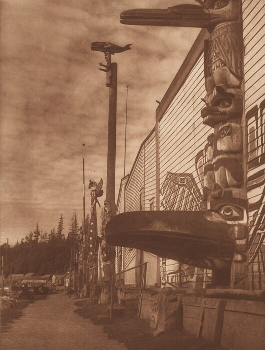 This is a photograph of a row of totem poles lined up alongside a building with a dirt path in front of them. The photograph captures several intricately carved totem poles, each distinct in design, featuring various animal motifs such as birds and sea creatures intricately carved and stacked vertically. The totem poles stand beside a large wooden building, the side of which is adorned with similar artistic designs. The sky above is overcast with scattered clouds, casting a soft light over the scene. To the left of the image, there is a progression of trees providing a natural backdrop, and in the distance, smaller structures can be observed, all contributing to the historical ambiance of the photograph.