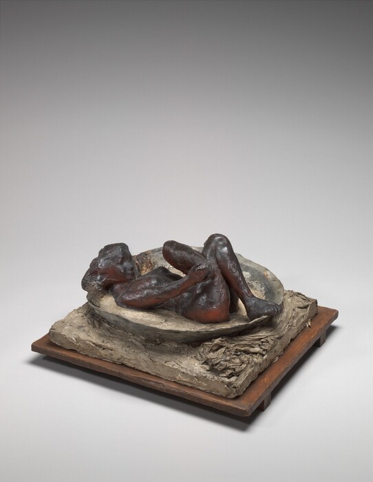 The sculpture depicts a nude woman lying in a shallow, oval basin with bent legs, grabbing one of her feet in her hand while the other foot rests on the rim of the basin. The sculpture appears to be made from a dense, earth-toned material, with the figure of the woman displaying a dark brown color that gradually shifts to a deep reddish hue, highlighting the contours and details of her body. The basin is positioned on a flat, textured, grayish-brown base on top of a wooden platform.