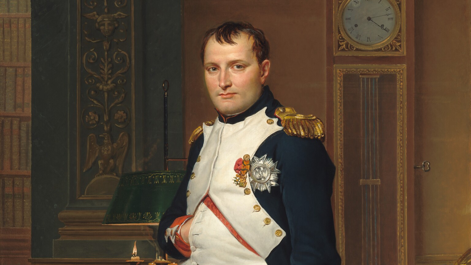 A man with pale peach skin and dark hair wears a military uniform and stands in front of a desk in this vertical portrait painting. He nearly fills the composition so seems close to us, and he looks directly at us. His body is angled slightly to our left and he tucks his right hand, on our left, flat against his chest between the buttons of his jacket. His navy-blue waistcoat is white along the front where it is fastened with brass buttons along his chest. The jacket has red cuffs, gold epaulets on the shoulders, and three medals affixed to the chest. White britches end just below the knee, and white stockings covering his calves are wrinkled at the ankle above black shoes with brass buckles. A candle burns low in a lamp on an ornately carved and gilded desk behind the man. Books and papers are piled on the desk to our right. More papers and a thin sword rest on a chair in front of the desk to our right. The chair is also carved and gilded, and is upholstered with scarlet-red fabric decorated with gold bees. The legs of the chair push back the forest-green carpet underfoot. A tall clock stands on the wall opposite us and reads 4:13. A few capital letters are written on a scroll of paper on the chair, “COD.” The artist’s name is also written as if printed on a scroll of paper on the floor behind the desk to our left: “LVD.CI.DAVID OPVS 1812.”