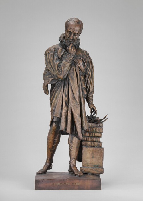 The sculpture depicts a man in a historically styled garment with a contemplative expression. He is leaning slightly with his left arm bent and right hand resting on a stack of books tied together with a strap. The sculpture is made of bronze and stands on a base inscribed with the name "Ambroise Paré."