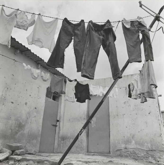 The image shows clothes hanging on a line in an outdoor setting. Various types of clothing are strung across a clothesline. The background features two weathered doors and aged walls. In the foreground, a wooden pole supports the clothesline, and the ground is uneven with scattered stones. The cloudy sky contrasts with the monochrome palette of the scene. The image conveys simplicity and the domestic ritual of drying laundry outdoors.
