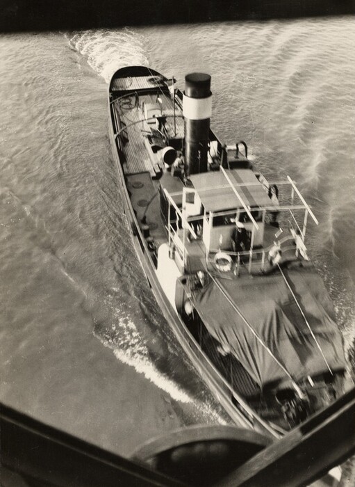 The image shows a boat navigating through water. The boat is angled diagonally across the image, with its bow pointing toward the top left corner. The hull is mostly dark with some lighter sections, while the smokestack stands out with its black and white stripes. A small wake follows the boat, creating ripples on the water. The photo is in black and white, with various shades of gray.