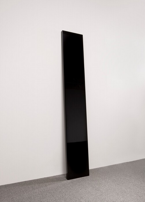 A tall, rectangular, highly polished black panel leans against a white wall. The shiny surface reflects the room, including the gray carpet.
