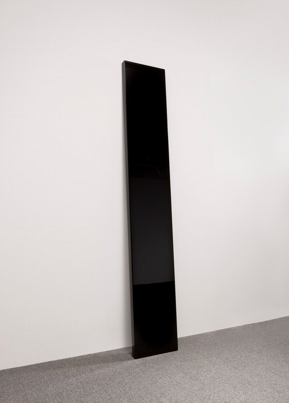 A tall, rectangular, highly polished black panel leans against a white wall. The shiny surface reflects the room, including the gray carpet.
