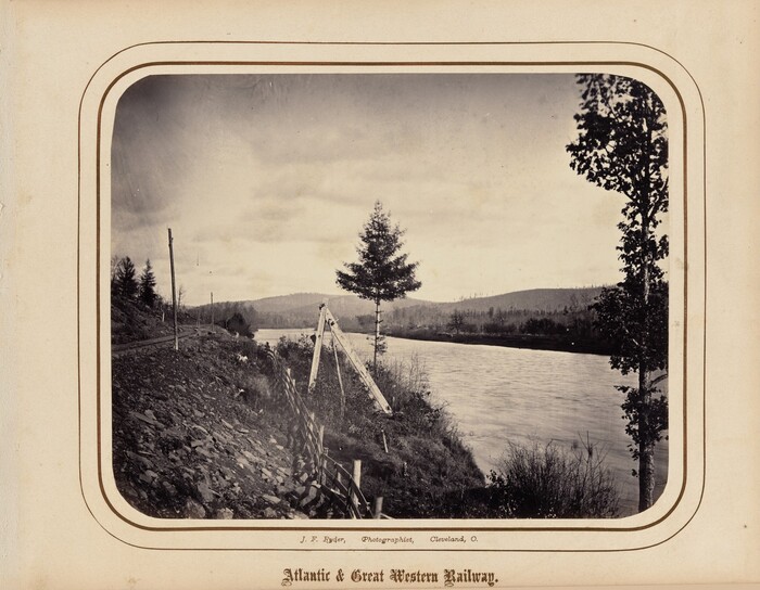 The image shows a river spanning from right to left, with a low horizon featuring rolling hills in the distance. On the left side, a rocky slope descends toward the river, with a railway track beside it. A pole is in the foreground, along with a bridge or trestle structure. The scene has a sepia tone, creating a historical feel. Trees on the right lean towards the water, blending into the sky. The photograph's tones play between the darker land and trees and the lighter sky and water.
