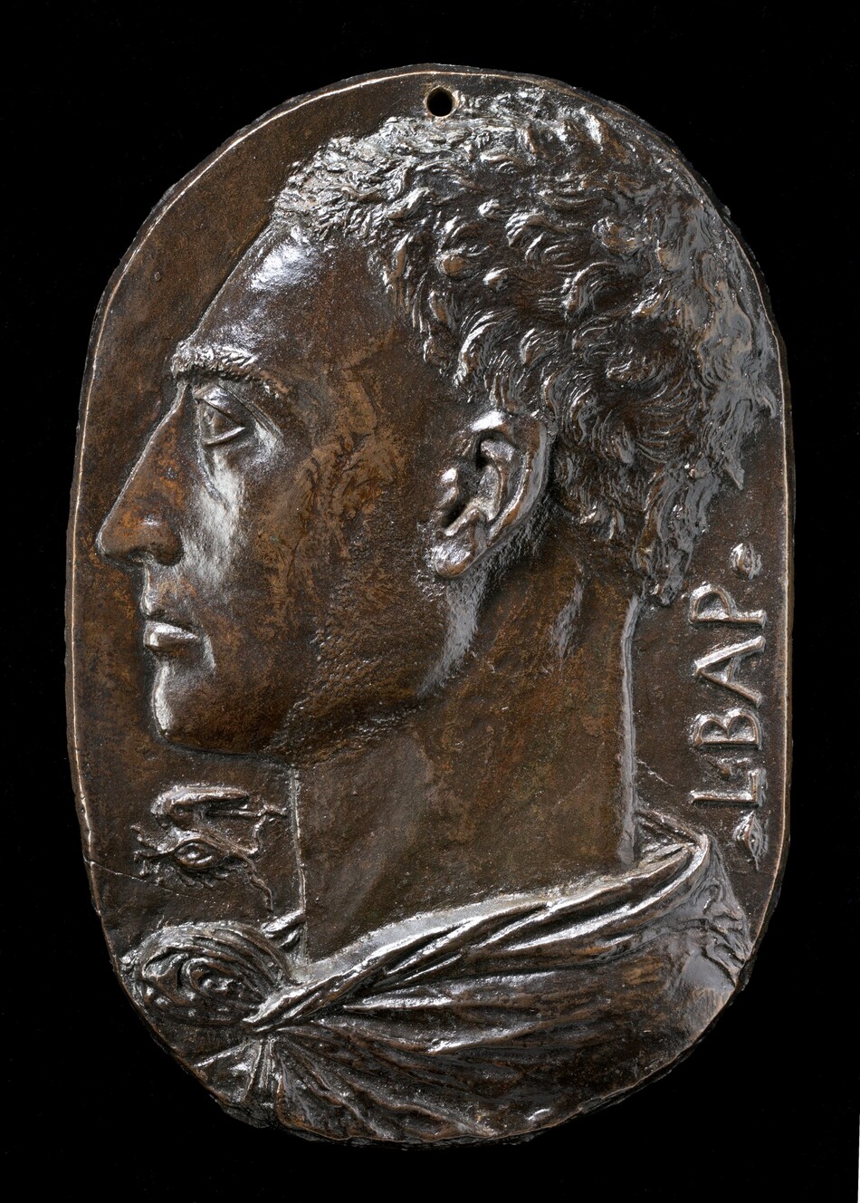 This bronze, oval medallion shows the profile of a man facing our left, his head and neck filling the space. He has short wavy hair and a long, pointed nose. He looks steadily into the distance to our left, and his lips are closed. His garment wraps around the back of his neck and is knotted at the base of his throat. The capital letters “L BAP” run vertically up near the edge of the medallion in the space behind his neck to our right. A symbol under his chin to our left is made up of a wing arched over a stylized eye. Light glints on the deep brown bronze surface, especially on the front of the chin, cheeks, and forehead, and in his choppy hair.