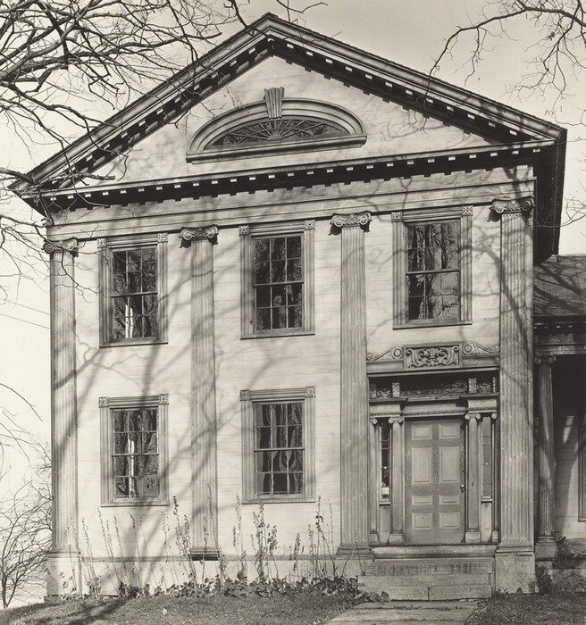 The image shows a classical-style building facade. It includes a grand building with a portico supported by four large Corinthian columns. The facade is symmetrical, with evenly spaced windows that have multiple panes, and an ornate doorway with decorative elements above it. An arched window is located above the entry door. The building is framed by leafless tree branches, casting shadows on its surface. There is sparse vegetation on the ground, suggesting minimalist garden landscaping.