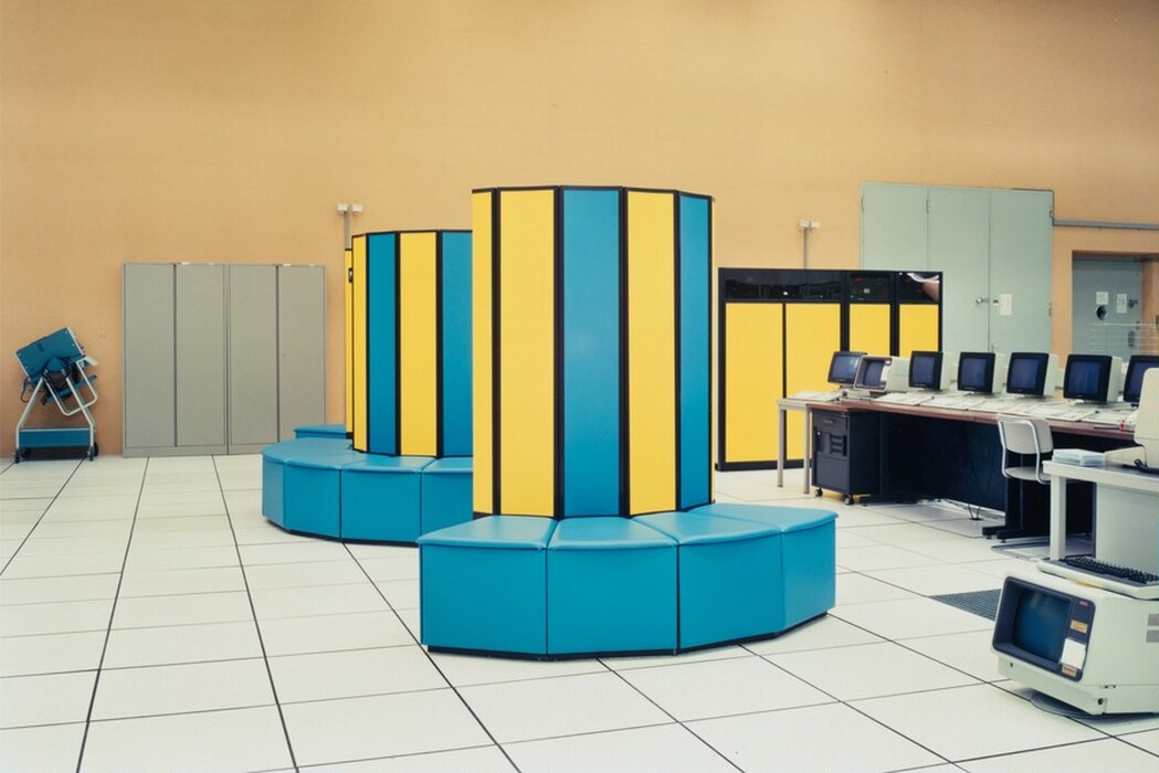 The image shows a computer room with large-scale computing equipment. The room has a light-colored tiled floor and beige walls. Two large cylindrical computer units with yellow and blue vertical panels are in the room. A row of vintage computer terminals is set up on a desk to the right, with their screens facing inward and connected by cables. Additional smaller computer units are on tables throughout the room. The design and arrangement suggest a focus on computing and data processing in the working environment.