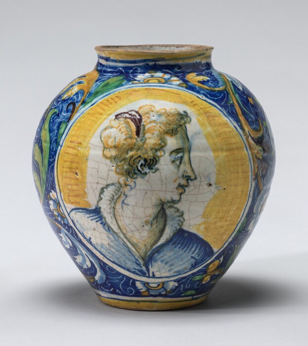 This is a photograph of a decorative ceramic jar with a portrait. The jar is adorned with vibrant colors featuring a central circular medallion depicting a profile portrait. The portrait is done in a classical style, with curly hair, an elaborate ruff collar, and delicate features facing left. The background of the medallion is styled in a warm yellow tone. Surrounding this central image, the jar has intricate patterns in blue, yellow, and green, incorporating leaves and floral motifs. The rounded shape of the jar, along with the rich colors and artistic glaze work, suggest an appreciation for classical art design. The craftsmanship and detail suggest the piece could be from the period of Renaissance ceramics.
