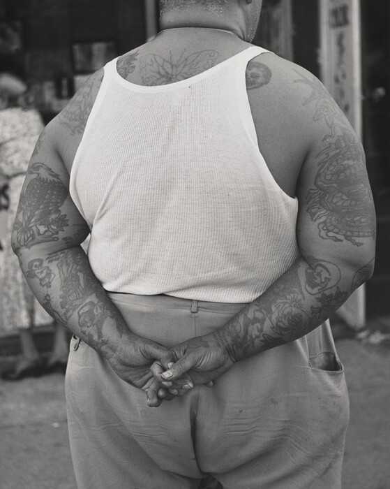 The image shows the back of a person's torso and part of their lower body. The individual is standing with their back to the camera, arms crossed behind their back. Their sleeveless white undershirt reveals both arms, which are heavily tattooed with various designs. They have short hair and wear khaki pants, with a thin chain around their neck. In the background, there is a partially visible figure in a floral dress and a hat, standing next to a wall with indistinct posters or signs hanging on it. The setting appears to be an outdoor urban area with a concrete surface.