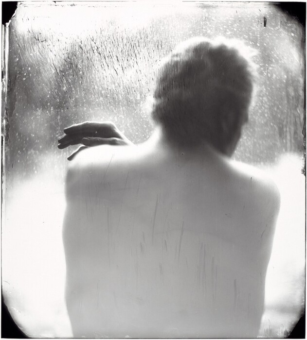 The image shows the back of a person from shoulders upwards, including part of their head. The person is positioned with their back slightly turned away from the camera, and their arm is raised to rest on their opposite shoulder. The facial features of the person are not visible, but the hair appears to be light in color and is softly styled or tousled. The person's skin is bare with no visible clothing. The background consists of a foggy or misted surface with droplets, possibly a window or glass pane, and there is a soft light illuminating the scene, enhancing the texture.