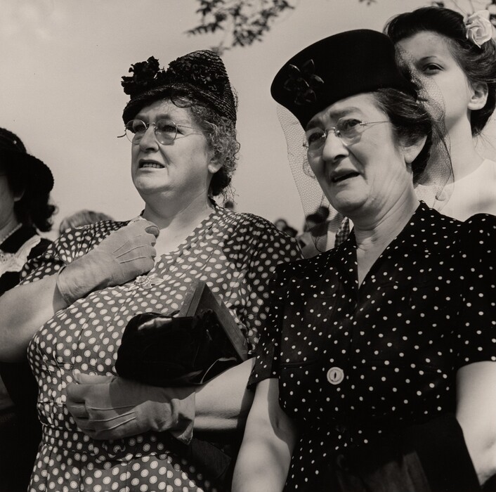 The image shows two women from their waists upwards, standing side by side. The woman on the left has curly hair adorned with a decorative hat featuring flowers, glasses, a light-colored polka-dotted dress, gloves, and a handbag. The woman on the right has her hair tucked under a decorative hat with a veil, glasses, a dark polka-dotted dress, and a collar. A partial view of a third person is visible on the right side. The background includes a clear sky with some tree branches partially visible.