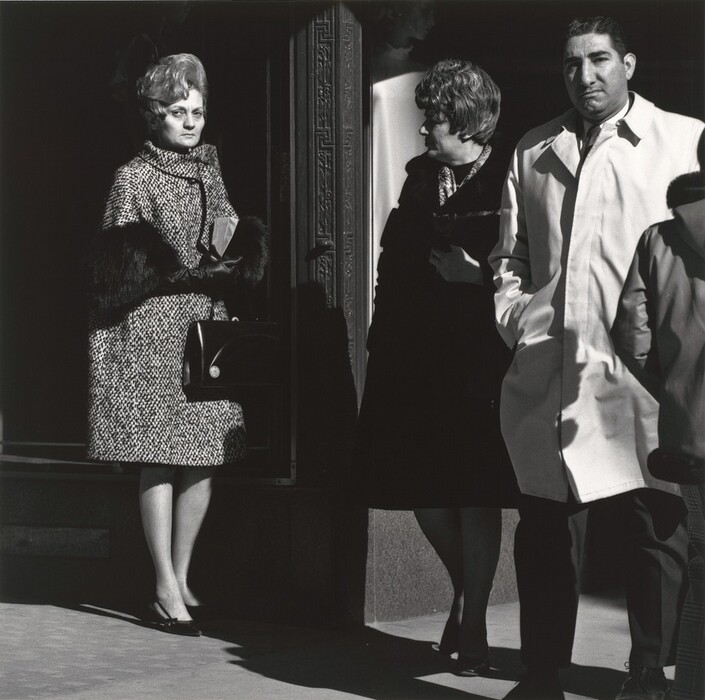 The image shows three individuals partially, with their full bodies visible up to their knees. The woman on the left is standing in an open doorway and is positioned at an angle, looking towards the camera. She has styled hair that appears voluminous and wavy. She wears a textured coat with dark fur cuffs, gloves, and holds a black handbag. Adjacent to her is another woman facing right, engaged with the man to her side. Her hair is shorter and possibly curly or wavy, and she is dressed in a dark outfit with a scarf. The man next to her stands facing forward, wearing a light-colored overcoat. The background features a structural design with ornate detailing around the doorway, and the setting seems to be an urban environment on a bright day, suggested by the sharp shadows.