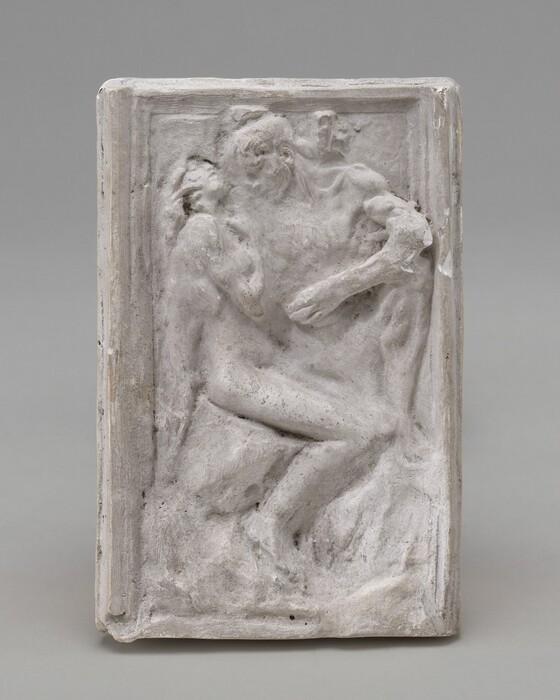 The sculpture features a relief of two intertwined human figures on a rectangular slab with a raised border. The material is a pale, stone-like substance. The figures are highly textured, depicting musculature and contours. The sculpture conveys movement and interaction, with the figures merging within the frame.