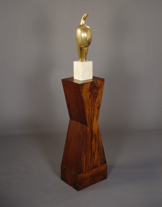 A polished bronze, free-standing sculpture of an abstracted bird stands on a marble plinth set on a tall wood base. The bird form is created with an oblong, egg-shaped body. The head curves up like a hook and the end parts as if in a beak. The wings are tucked against the body. The bronze sculpture sits on a cream-white, tall marble cube. Shaped like an angular hourglass, the wood base is almost twice as high as the bronze sculpture and marble cube combined.