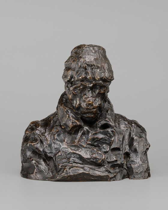 This sculpture is a bust of a person wearing a cap and a thick, crumpled overcoat. The person appears to have their head slightly bowed, and they seem to have deep set eyes and a long nose, although their facial features are hard to identify. The sculpture is made of a dark material that has a rough and uneven texture with deep grooves.