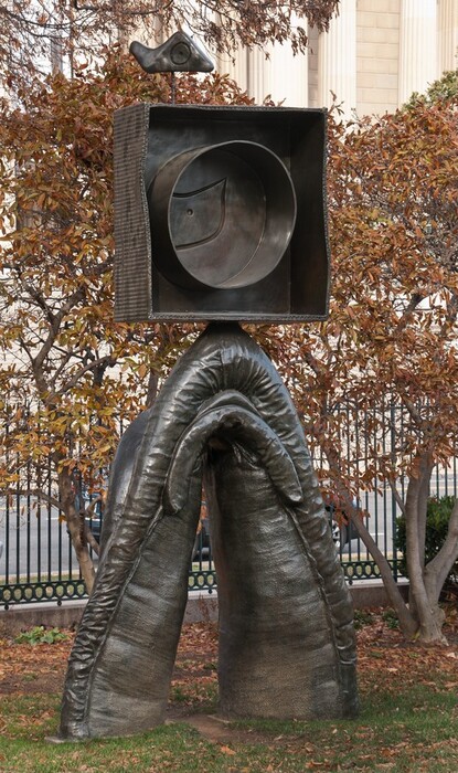 As tall as the spindly trees behind it, a bronze, abstract, free-standing sculpture is made up of a circle set within a shallow box, which is balanced on the tip of an upside-down, thick-legged, V shape. The textured, lumpy legs taper as they move up. A sinewy rope-like texture runs, like a hem, along the outside of the pointed arch. Another form, like a lip, lines the inner edge where the legs pinch together at the top of the arch. The box balanced on the apex is nearly as wide as the legs. The box has a projecting rim, and is corrugated along the outer surfaces and the front edge, as if it had been cut down from a cardboard box. The circle centered in the box also has a projecting rim, like a tall cake pan. Within the circle, a shape like the wing of a bird appears to have been incised. A curving, bowtie-like shape protruding on a rod from the top of the box is incised with a dot at the center of another circle.