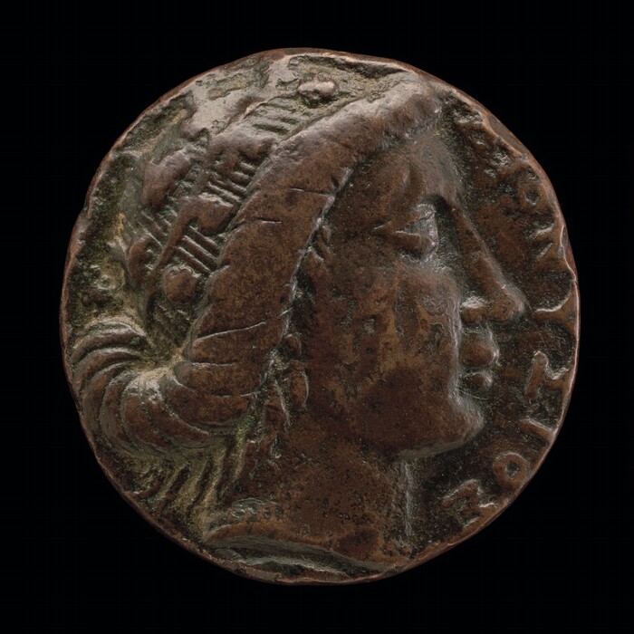 The image depicts a round coin-like sculpture with a human face profile, possibly of a historical or mythological figure. The sculpture's subject wears a headpiece with a braided or draped element behind the head. The material looks like brown metal, possibly bronze, with a visible patina and texture. The face is in profile view, facing right, with detailed features. The coin includes inscriptions around the edge, with some parts appearing worn or obscured, featuring a combination of letters and symbols.