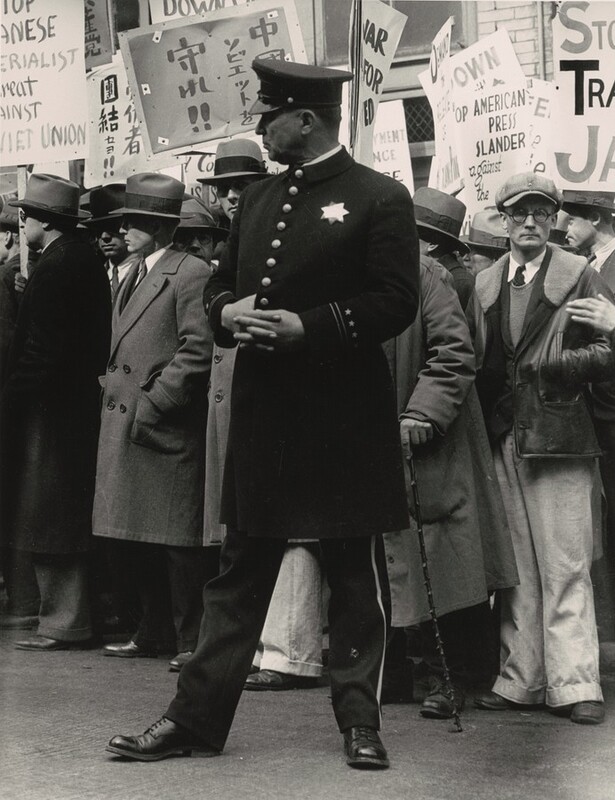 A uniformed policeman standing in front of a dense crowd of men holding protest signs in English and Japanese nearly fills the height of this vertical black and white photograph. The policeman either has a ruddy complexion or his face might be in shadow. He stands with his body facing us but he turns his head to our left in profile. His hands are crossed over his chest and one thumb is hooked into his jacket between two of the eight brass buttons down the front. A seven-pointed star reflects light on his chest and his flat-topped hat has a short, shiny brim angled down over his eyes. The jacket comes to his knees and his pants are lined with gold or other light material down the side. His feet are widely planted, and he wears dark shoes. The men in the crowd behind appear to be light skinned. Many wear button-down, collared shirts with ties tucked into long coats. Many also wear fedora hats and look off to our left, the same direction as the policeman. One man to our right wearing a floppy cap and glasses looks out at us with his hands in his jacket pockets. Some of the legible English words on the protest signs read “UNION,” “AMERICAN PRESS SLANDER against the,” and “JA.”