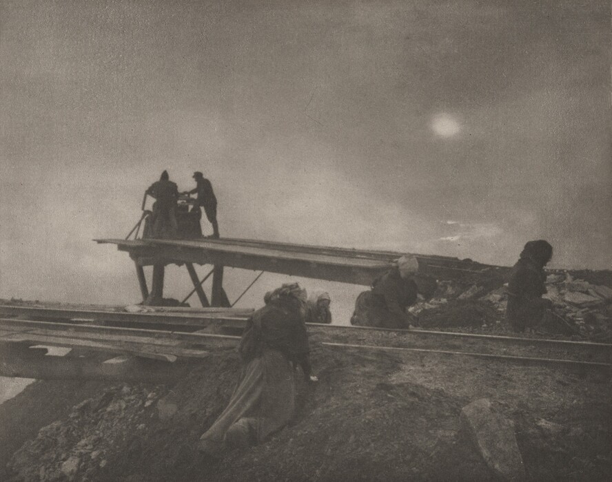 This is a photograph of an industrial landscape with figures engaged in construction work on a mound of earth beneath a large, wooden structure. The scene is captured in black-and-white, giving it a historical or vintage feel. The photograph showcases an elevated platform in the background with two silhouetted figures standing. The foreground presents rugged terrain with loose earth and rocks, where additional figures appear to be engaged in manual labor. The sky above is cloudy with diffused sunlight casting soft illumination over the workers and construction scene. The composition highlights hard labor and industrialization.