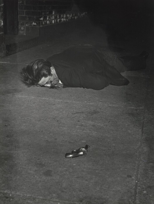This is a photograph of a crime scene. The image shows a person lying face down on the pavement, with blood nearby and a handgun on the concrete surface. A brick wall runs along the left side of the scene. The lighting is stark, with high contrast and deep shadows.