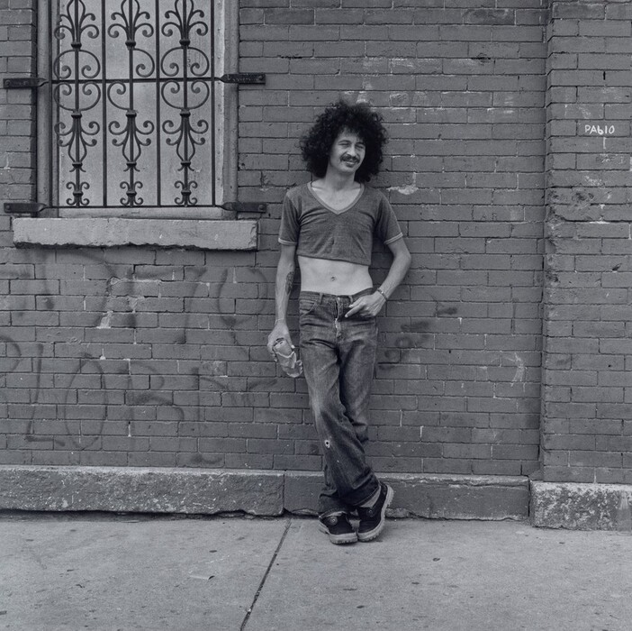 The image shows a person leaning against a brick wall on a sidewalk. The person has a relaxed posture, with their left elbow on the wall and right hand in their pocket. They have curly hair, a slight mustache, and a smile. They wear a cropped V-neck T-shirt, jeans with a hole in one knee, and dark clogs. They hold a crumpled brown paper bag in their left hand and have a small wristband on their left wrist. The background includes a brick wall with an iron window grill, faded graffiti, and the word "PABLO."