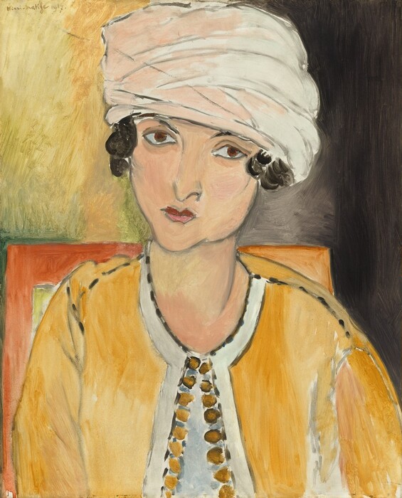 Shown from the chest up, a woman with a pale, peachy skin wears a turban and a goldenrod-yellow jacket in this stylized, vertical portrait painting. Many of the features and forms are outlined in gray and filled in with areas of mottled color so brushstrokes are visible. The woman’s shoulders face us, and she tips her head a little to our right. One brown eye, to our left, seems to look at us, and the other might look off over our right shoulder. Her nose is indicated with a wavy line for a pointed tip, and her small pink lips are closed. She has a square jaw and a narrow chin. Black hair curls over her ears. The rest of her hair is covered by a turban, which touches the top edge of the composition and is pale pink to our left and ivory white to our right. Her yellow garment is fastened at her throat and has rows of darker gold circles, perhaps buttons or other trim, down the open front over a white shirt. The papaya-orange corners of a square-backed chair is behind her shoulders, about halfway up the picture. The background is streaked with laurel green, flax, and lemonade yellow to our left, and deepens to dark, pewter gray to our right. The artist signed and dated the painting with olive-green script in the upper left corner: “Henri-Matisse 1917.”