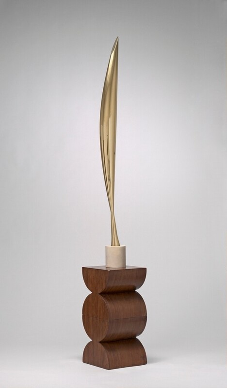 The top section of this abstract sculpture is made up of a vertical, elongated form cast in gleaming brass that swells gently at the center and tapers to a point at either end. Near the bottom point, the form flares out slightly to make a tall, conical foot. This sits atop a short, cylindrical, white limestone base on a wood pedestal. The pedestal is carved to look like a disc at the center flanked above and below by semicircles with the flat edges facing up and down. The sculpture is photographed against a pale gray background.