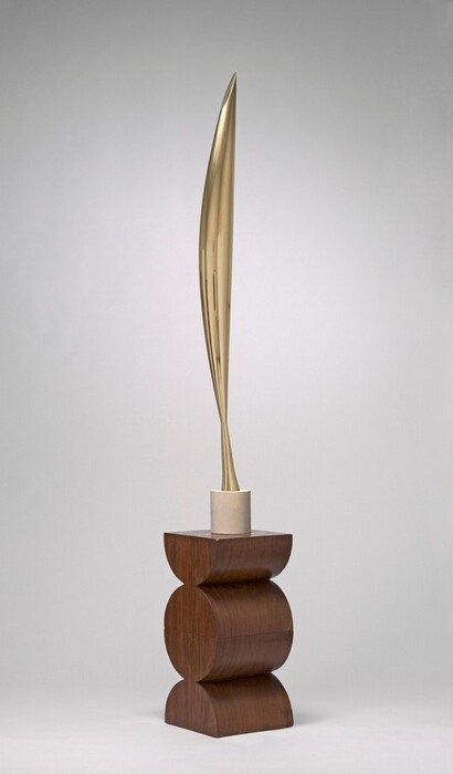 The top section of this abstract sculpture is made up of a vertical, elongated form cast in gleaming brass that swells gently at the center and tapers to a point at either end. Near the bottom point, the form flares out slightly to make a tall, conical foot. This sits atop a short, cylindrical, white limestone base on a wood pedestal. The pedestal is carved to look like a disc at the center flanked above and below by semicircles with the flat edges facing up and down. The sculpture is photographed against a pale gray background.