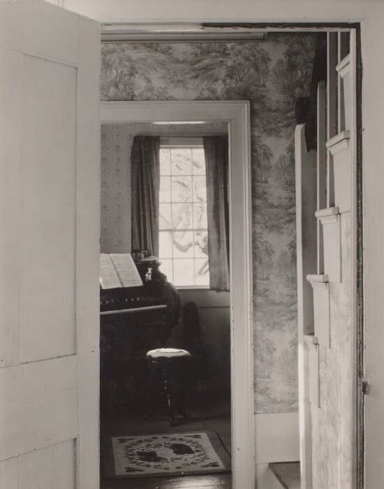 This is a photograph of an interior view through a series of doorways leading into a room with a musical instrument and sheet music. The image captures a hallway with an open door, beyond which is another room. The walls are adorned with patterned wallpaper featuring a pastoral scene with trees. A window in the far room lets in natural light, casting shadows through thin curtains. Near the window is a musical instrument with sheet music placed on top, and a cushioned seat is in front of it. A small rug with a decorative design lies on the floor. The photograph conveys a sense of depth and perspective through its composition of the receding doorways.