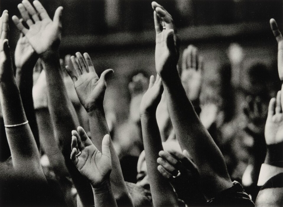 This is a photograph of raised hands with open palms. The black and white image captures several hands raised in different angles. The focus is on the hands, creating a sense of unity and motion. The composition shows a variety of positions and overlap of hands. Details like lines on palms and accessories like bracelets and rings can be seen, highlighted by the contrast. The background is blurred, emphasizing the hands in the foreground.