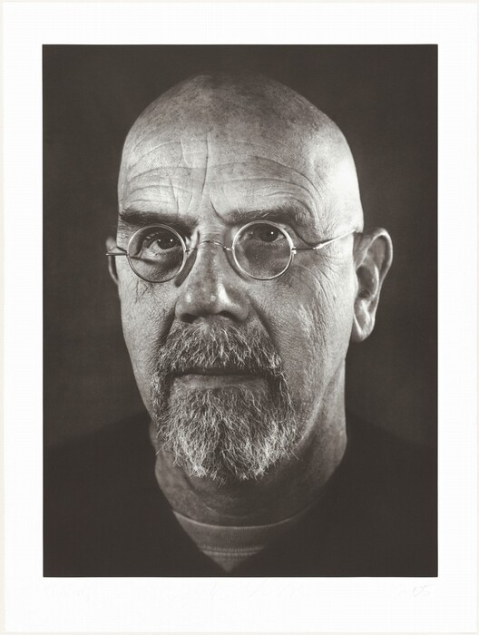 The image shows a close-up of a person's face, capturing only their head and upper portion of their shoulders. The person is facing forward towards the camera in an upright position. The individual has a prominent bald head with no visible hair on the scalp. They have round, wire-rimmed glasses that accentuate their eyes. The facial features include a strong, wide nose, defined wrinkles on the forehead, and a well-groomed, full beard and mustache, showing a mix of gray and white color. The person is wearing a dark-colored top, and the image does not reveal any jewelry or additional objects. The background is plain and dark, providing a strong contrast to highlight the person's features.