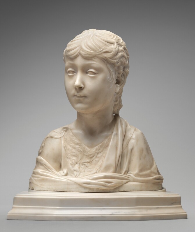 The head and shoulders of a young boy have been carved from white marble in this freestanding sculpture. In this photograph, the boy’s shoulders are square to us, and he turns his head to look off to our left. His wavy hair comes low on his forehead and curls along the back of his neck. He has slightly raised brows, and the irises of his eyes are lightly carved so the boy appears to look up under heavy lids. He has an oval face with smooth cheeks, a delicate nose, and his lips are closed. He wears a garment that appears to be fur under a more voluminous robe, which drapes over one shoulder and wraps around the chest. The sculpture ends in a straight, blunt line below the shoulders, and it sits on a stepped, white marble base. The background is pale gray.