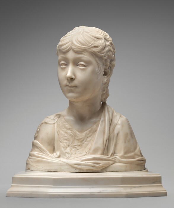 The head and shoulders of a young boy have been carved from white marble in this freestanding sculpture. In this photograph, the boy’s shoulders are square to us, and he turns his head to look off to our left. His wavy hair comes low on his forehead and curls along the back of his neck. He has slightly raised brows, and the irises of his eyes are lightly carved so the boy appears to look up under heavy lids. He has an oval face with smooth cheeks, a delicate nose, and his lips are closed. He wears a garment that appears to be fur under a more voluminous robe, which drapes over one shoulder and wraps around the chest. The sculpture ends in a straight, blunt line below the shoulders, and it sits on a stepped, white marble base. The background is pale gray.