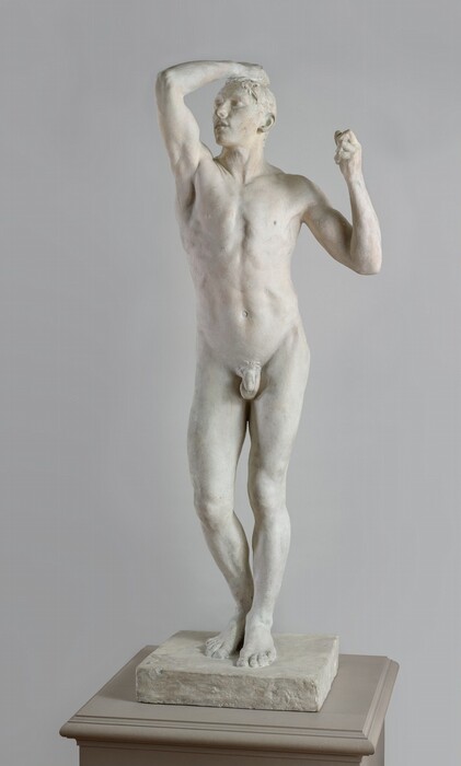 This free-standing, ivory-white plaster sculpture shows a nude, muscular, young man standing with his right hand, to our left, raised to the top of his head. In this photograph, his body faces us but his head turns slightly to our left toward the raised hand. He stands on a square base with his feet together and his right knee, on our left, slightly bent. That arm is raised in front of him with the elbow bent back so his hand grasps his short hair. His eyes and full mouth are closed. His other arm is also bent at the elbow at his side so that hand is at shoulder level, his fist curled. His face and arms have areas of straw yellow and flecks of caramel brown, presumably where the sculpture has been handled or worn.