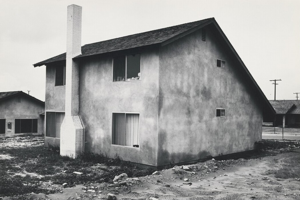 The image shows a ground-level point of view of a two-story building with a side view, featuring a large brick chimney extending from the ground to the roof. The horizon is low, with simple buildings and power lines visible against the cloudy sky. The house has a rough, textured exterior with minimal detailing, multiple rectangular windows, and a modernist aesthetic. The color palette is monochromatic due to the black and white photograph, emphasizing light and shadow contrasts on the building. Close to the building, there is rough land with stones and sparse grass leading up to the concrete base. In the distance, similar structures and utility poles are visible along a quiet residential street.