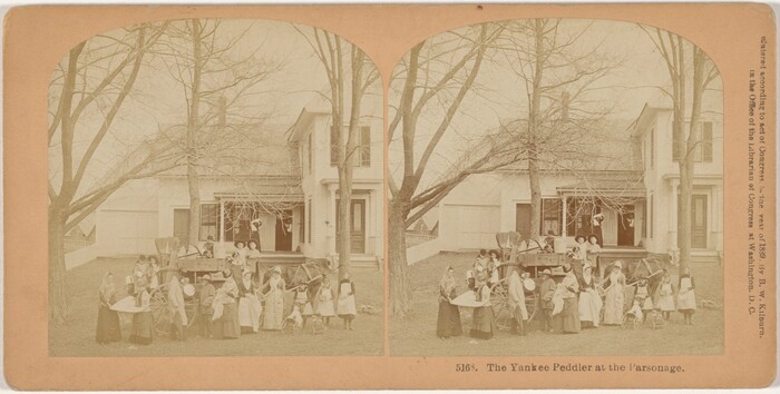 This is a photograph of a house with a group of people and a horse-drawn wagon in front of it. The image captures the facade of a large, two-story house with a porch and an adjacent wooden structure. In front of the house, a horse-drawn cart is parked with various items in it. Surrounding the wagon, a group of individuals are congregated. Two large, leafless trees frame the scene. This is a stereoscopic image.