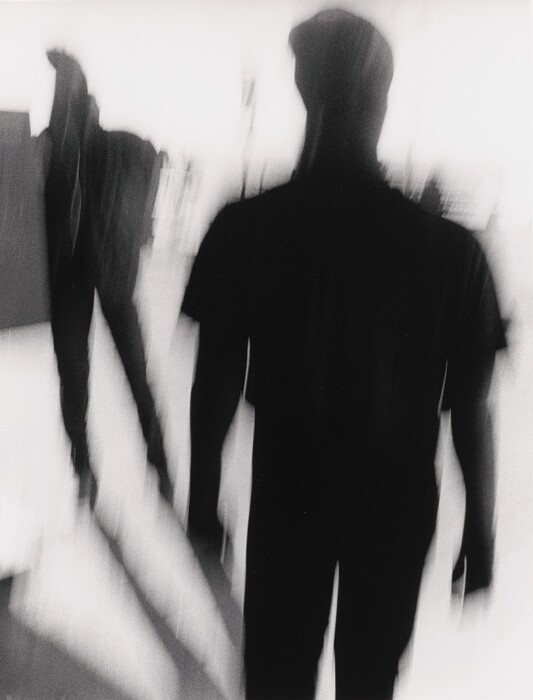 The image shows dark, blurred silhouettes with elongated and distorted shapes creating an abstract composition. The figures resemble human forms, suggesting motion. Their indistinct outlines and absence of detail give them a shadow-like quality, reminiscent of shadows or reflections in motion. The interplay of light and shadow, along with the dynamic lines, enhances the abstract nature of the scene.