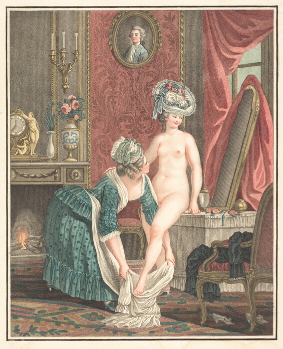 The image shows two women in an indoor setting. One woman is fully naked except for a decorative hat with flowers. The other woman, partially visible from the side, is helping the standing woman. The room contains a mirror, a plush chair with fabric, a side table with a vase of flowers, a clock, and candles. The wall has red-patterned wallpaper and an oval portrait of a man. There is a small fire and an ornate sconce.