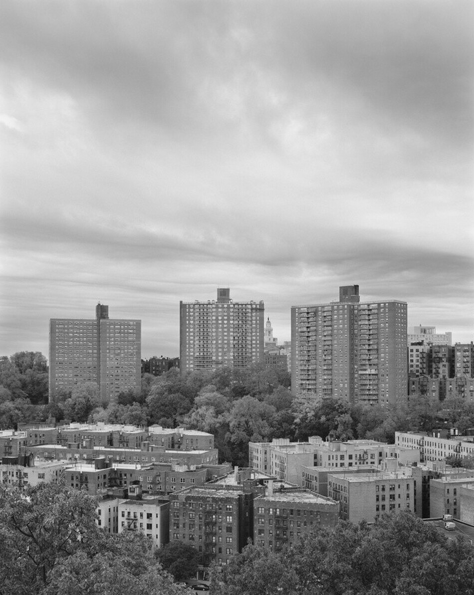 This grayscale photograph depicts a cityscape seen from an elevated viewpoint. Closest to us, there are several mid-rise apartment buildings with flat roofs and rectangular windows, densely arranged. Beyond these, a thick layer of trees creates a barrier between taller apartment towers. The sky above is vast and filled with dense, textured clouds, creating a dynamic and moody atmosphere.