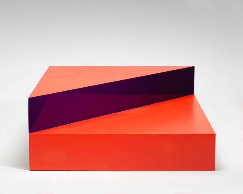 This abstract, free-standing sculpture is made up of a square, scarlet-red box like a platform topped with a triangle, half the size of the square. In this photograph, we look onto one side of the square base, and the triangle lines up with the half farther from us. The front face of the triangle is a reflective, amethyst-purple surface that reflects the square box beneath, and the top surface is coral orange. The backdrop behind the sculpture fades from light gray along the top of the photograph to white along the bottom.