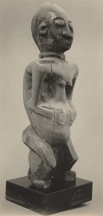 This is a grayscale photograph of a sculpture depicting a stylized human figure with an elongated head and round facial features, including closed eyes and a long nose. The figure is shown seated or crouching with their arms resting on their stomach, their back slightly curved. It is angled slightly to the right. The sculpture appears to be made from wood with a weathered appearance that includes visible cracks and a rough texture. It is mounted on a smooth, dark base. The background is plain gray.