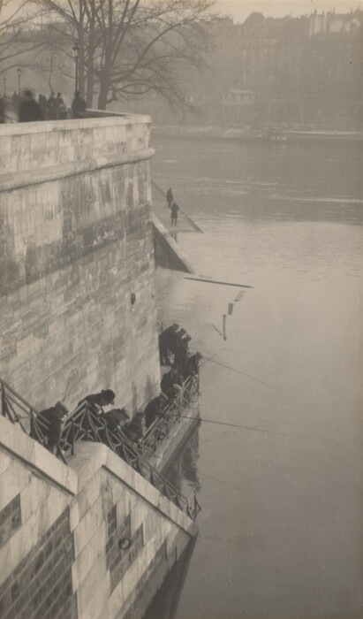 The image shows a top-down view of stone steps leading to a river, where people are fishing. Buildings line the riverbank, with trees in the background. The scene is monochromatic with various shades of gray, giving it a somber atmosphere. The texture is smooth, with detailed stone surfaces. The water appears calm. City buildings in the distance are partially obscured by mist.